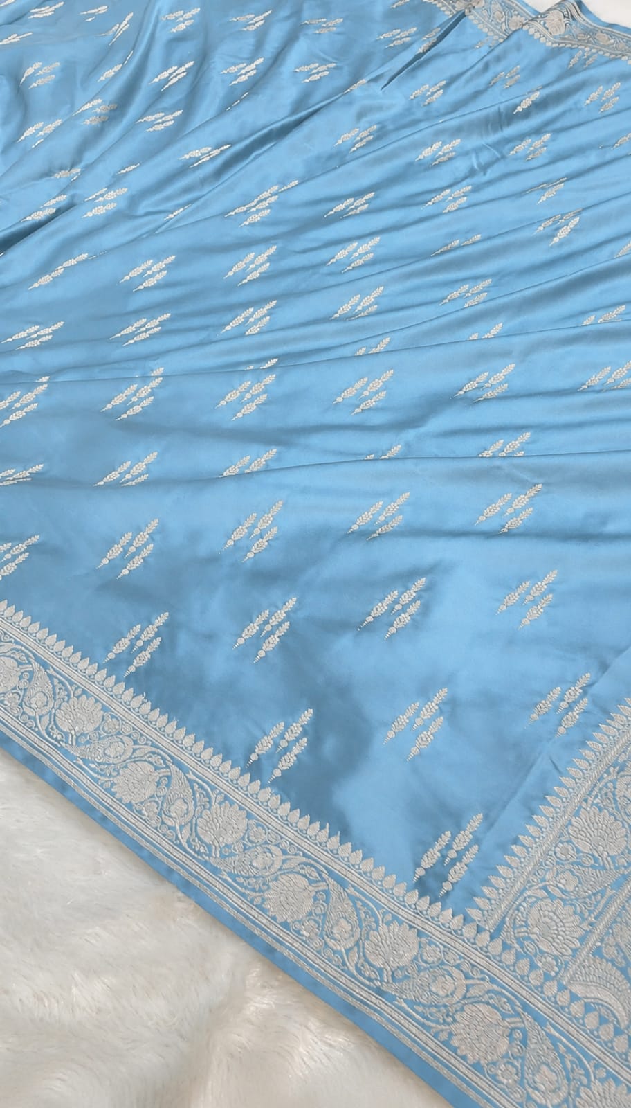 A light blue fabric garment or textile laid flat, featuring small repeating white embroidered motifs and a wide ornate white border along one edge, photographed on a white textured surface.