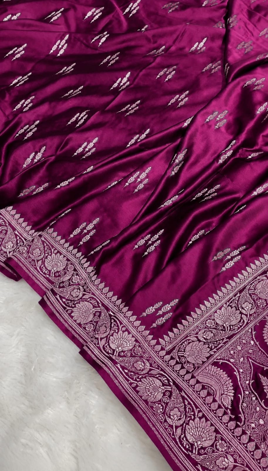 A close-up of a rich magenta purple silk saree with intricate silver zari embroidery and a detailed paisley border, laid over a white textured surface.