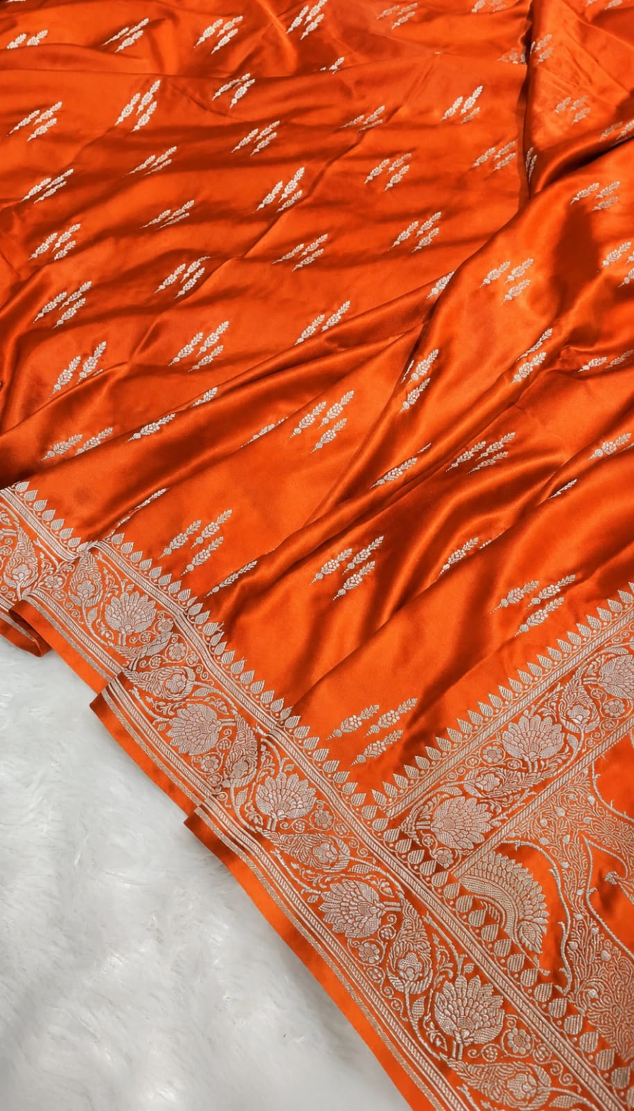 An orange silk saree with intricate silver Banarasi motifs and a detailed silver border, laid out on a white textured surface.