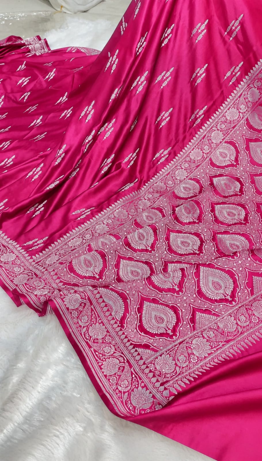 A close-up of a bright pink fabric garment, likely a sari or dress, featuring intricate silver paisley and floral border embroidery and small silver motifs throughout.