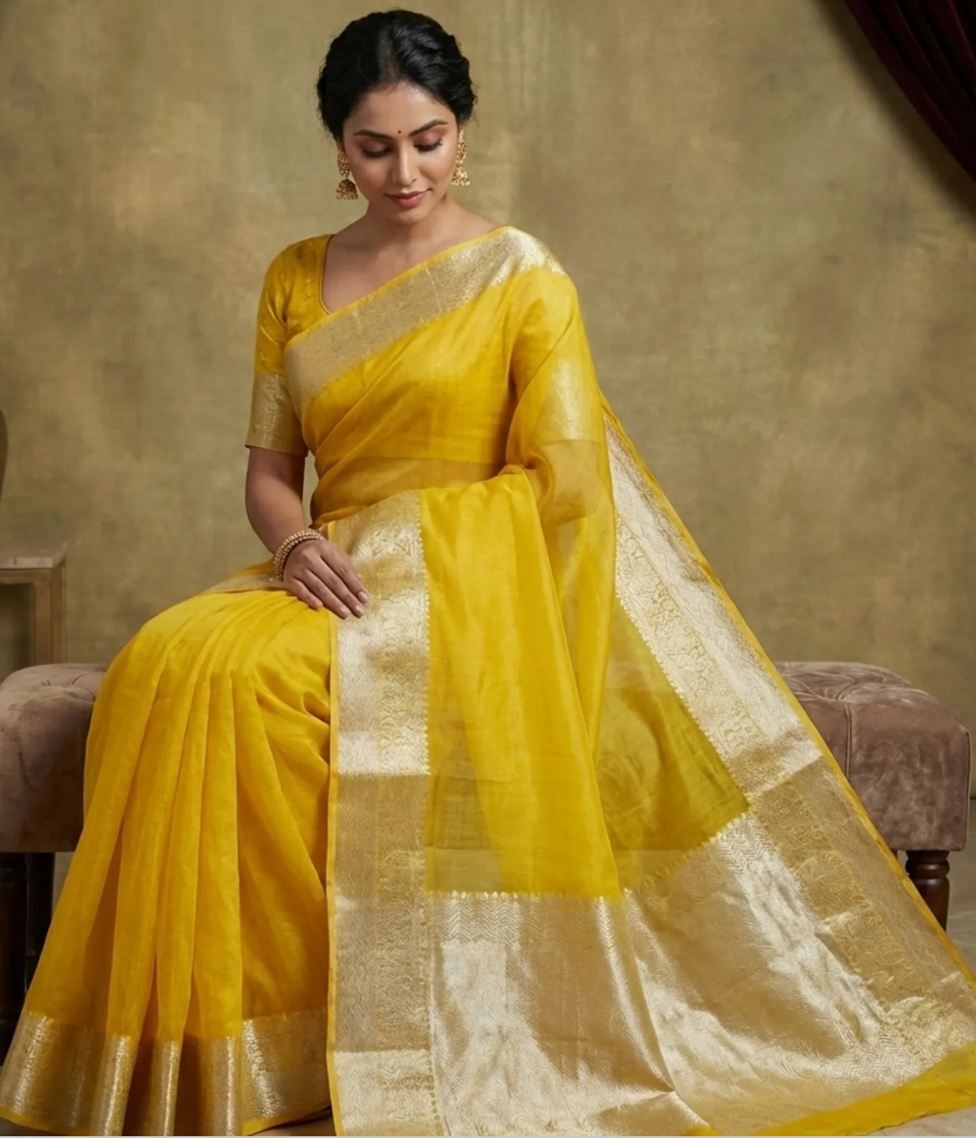 A woman seated on a cushioned bench wearing a bright yellow saree with a wide metallic gold border and matching blouse, photographed against a warm beige studio backdrop.