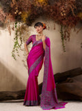 Woman in a pink saree with intricate patterns standing against a natural backdrop.