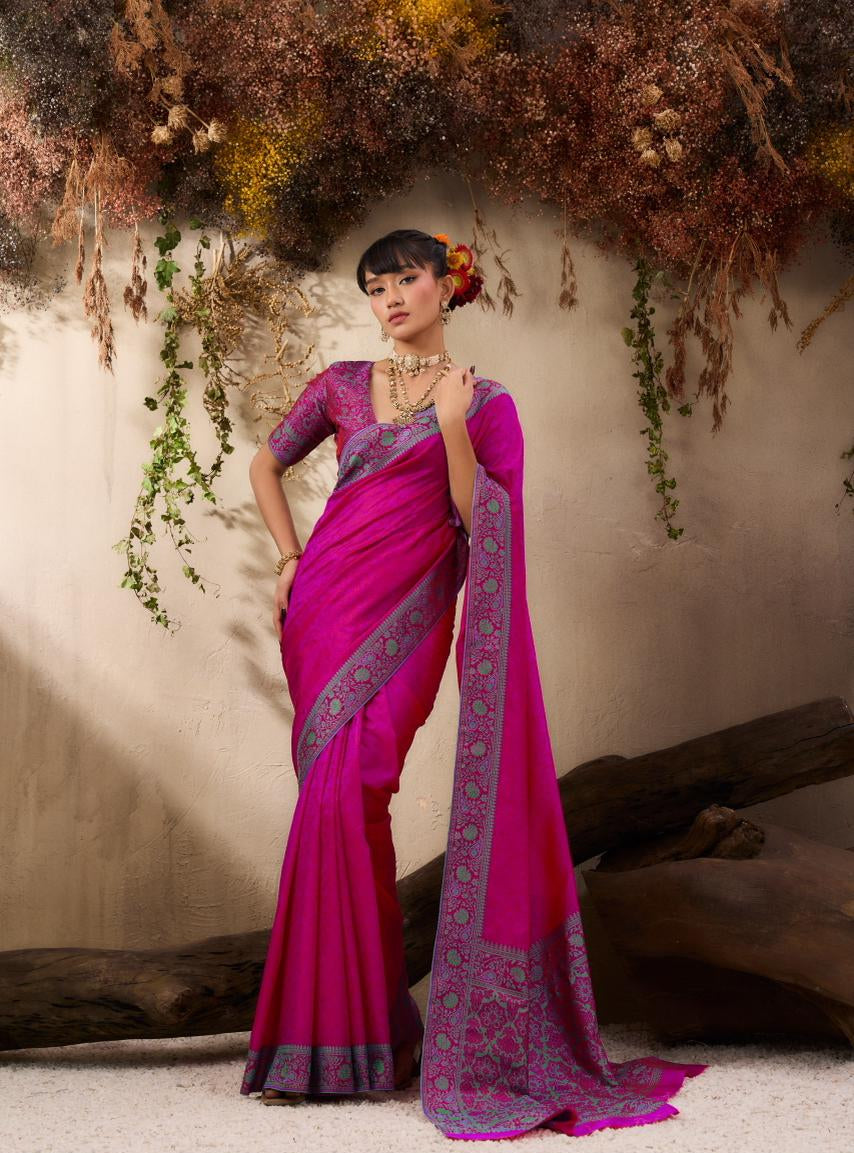 Woman in a pink saree with intricate patterns standing against a natural backdrop.