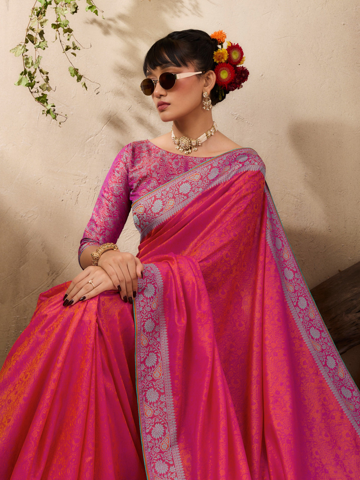 A woman wearing a bright pink and magenta saree with ornate light blue borders, seated against a textured beige wall with decorative flowers in her hair.
