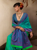 A woman wearing a vibrant teal and purple patterned saree with a matching blouse, styled with traditional jewelry and a floral hair accessory, seated against a textured beige wall.