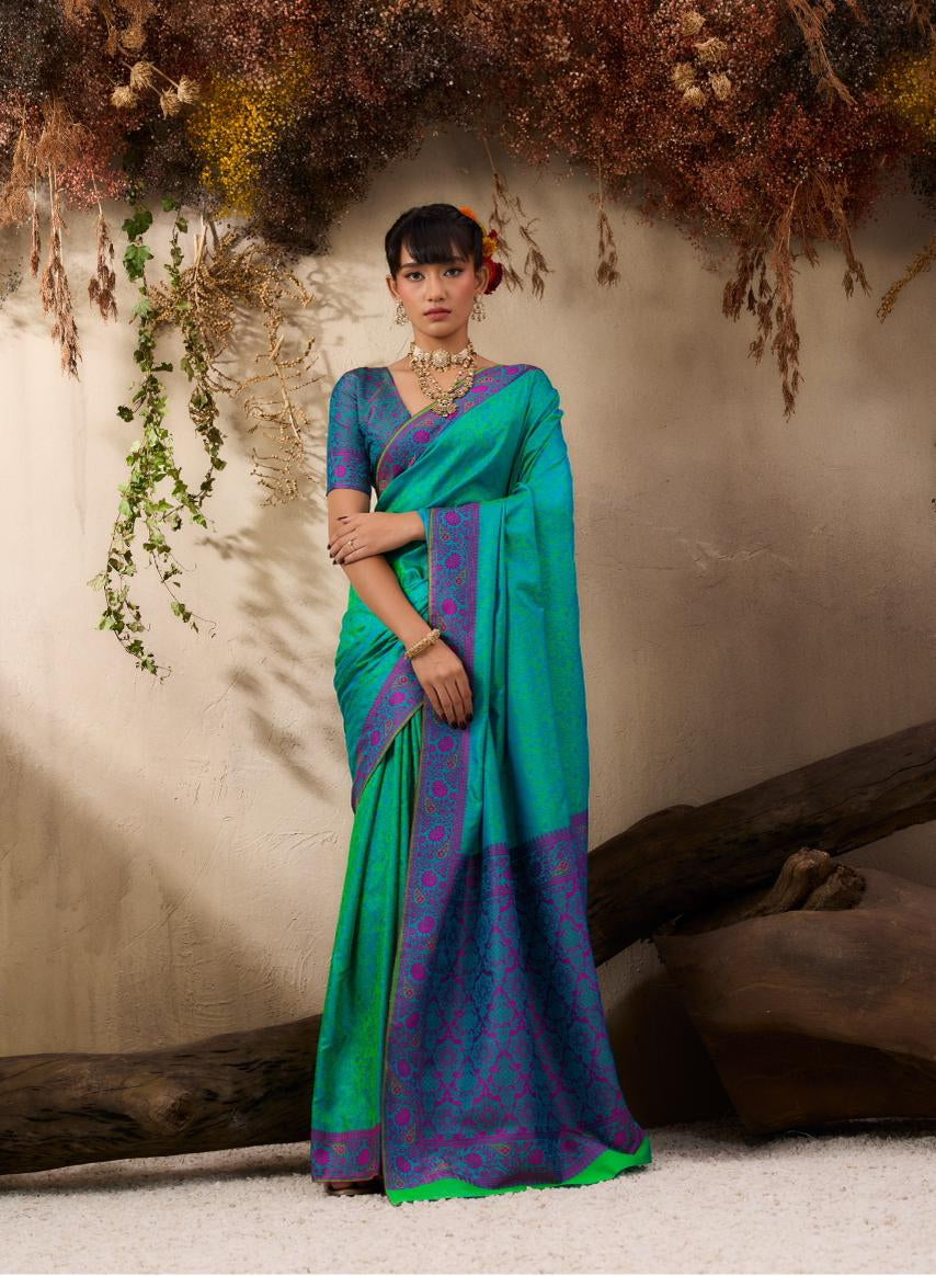 A woman models a Kanjivaram Tanchoi saree in a styled studio setting with dried floral decor. The saree features a vibrant teal green body with a purple and blue patterned border and a solid purple blouse.