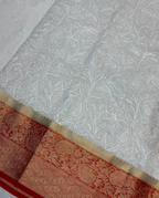 White embroidered fabric with intricate floral and paisley patterns, featuring a decorative border in beige and red with metallic woven detailing.