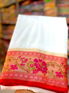 A folded white fabric item, likely a saree pallu or dupatta, held up by a hand in a shop. The bottom edge features a wide border with pink floral embroidery and gold zari detailing on a red base.