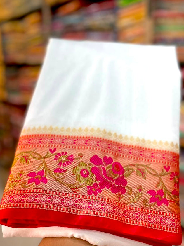A folded white fabric item, likely a saree pallu or dupatta, held up by a hand in a shop. The bottom edge features a wide border with pink floral embroidery and gold zari detailing on a red base.