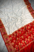 A white silk saree with a rich red border featuring intricate gold and pink floral patterns, draped on a gray marble surface.