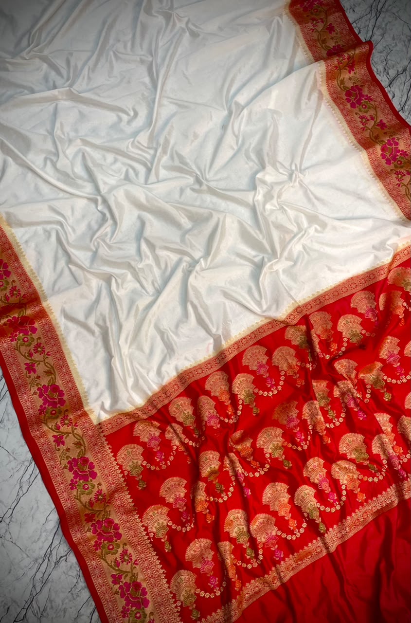 A white silk saree with a rich red border featuring intricate gold and pink floral patterns, draped on a gray marble surface.