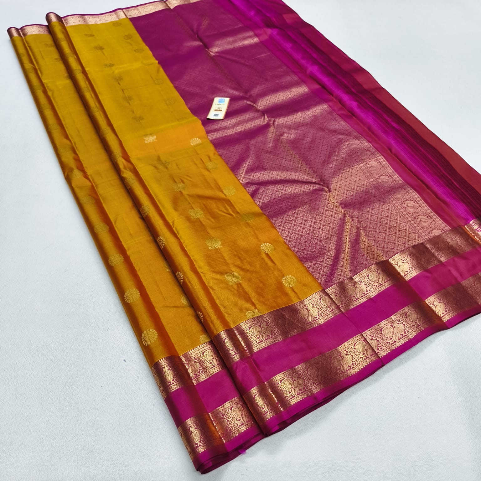 A folded silk saree with a mustard yellow body, a magenta-pink border, and intricate gold zari woven patterns, displayed on a light gray background.