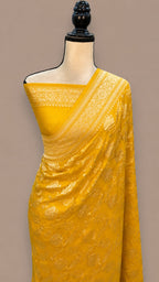 A bright yellow saree with intricate gold zari embroidery, draped on a mannequin torso against a solid taupe background.