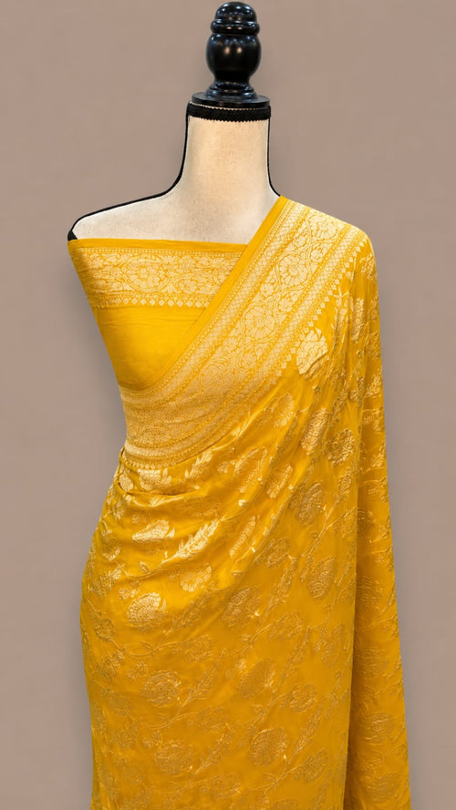 A bright yellow saree with intricate gold zari embroidery, draped on a mannequin torso against a solid taupe background.