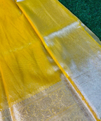 A close-up of a yellow sheer fabric, likely a sari or dupatta, featuring a wide silver metallic border with intricate woven patterns, laid out on a dark green textured surface.